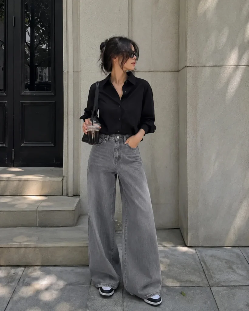 woman wearing a classic black button-up shirt tucked into high-waisted gray wide-leg jeans. The dark structured top creates a sleek, sophisticated contrast with the relaxed washed denim, giving the outfit a modern city-chic feel. 
