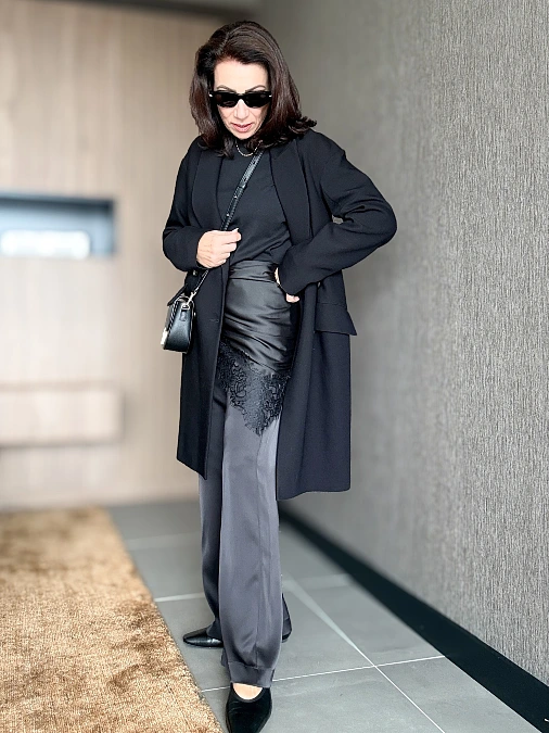 Woman wearing a long black tailored coat, black top, satin-look wrap layer with lace trim, gray wide-leg trousers, black shoes, and sunglasses.