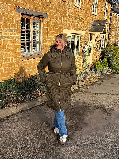 Woman wearing a knee-length olive green quilted hooded parka, blue straight-leg jeans, and casual sneakers outside a stone building.