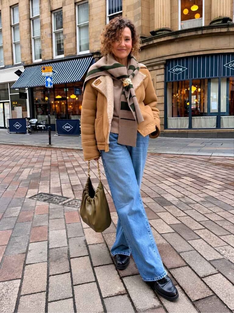 Woman wearing a tan shearling-lined jacket, striped scarf, taupe sweater, wide-leg blue jeans, black loafers, and an olive-green handbag on a city street.