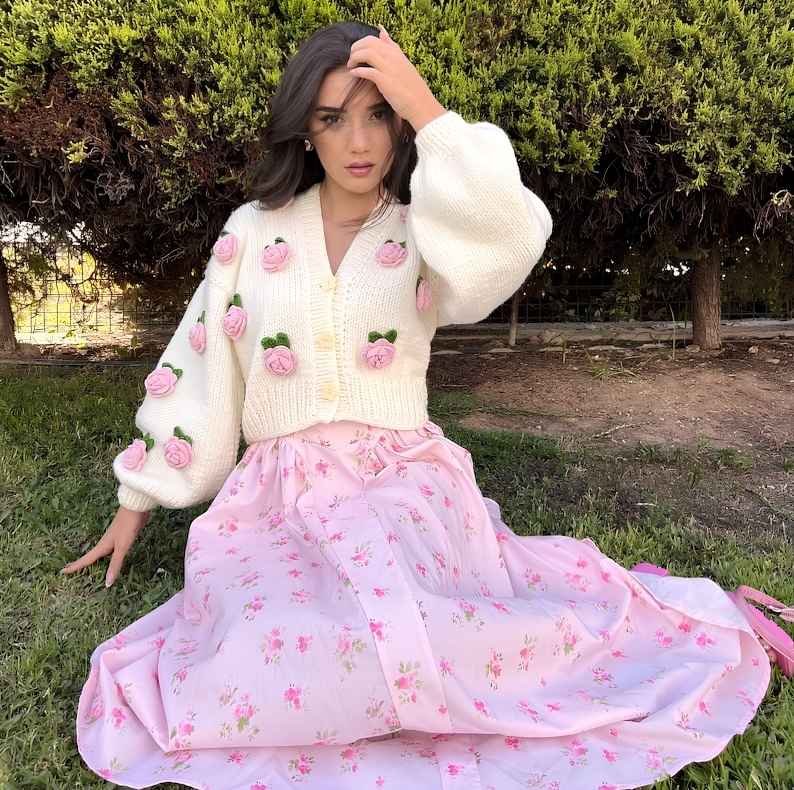 Woman wearing a cream crochet cardigan with pink rose appliqués and balloon sleeves, paired with a flowing pastel pink floral maxi skirt outdoors.