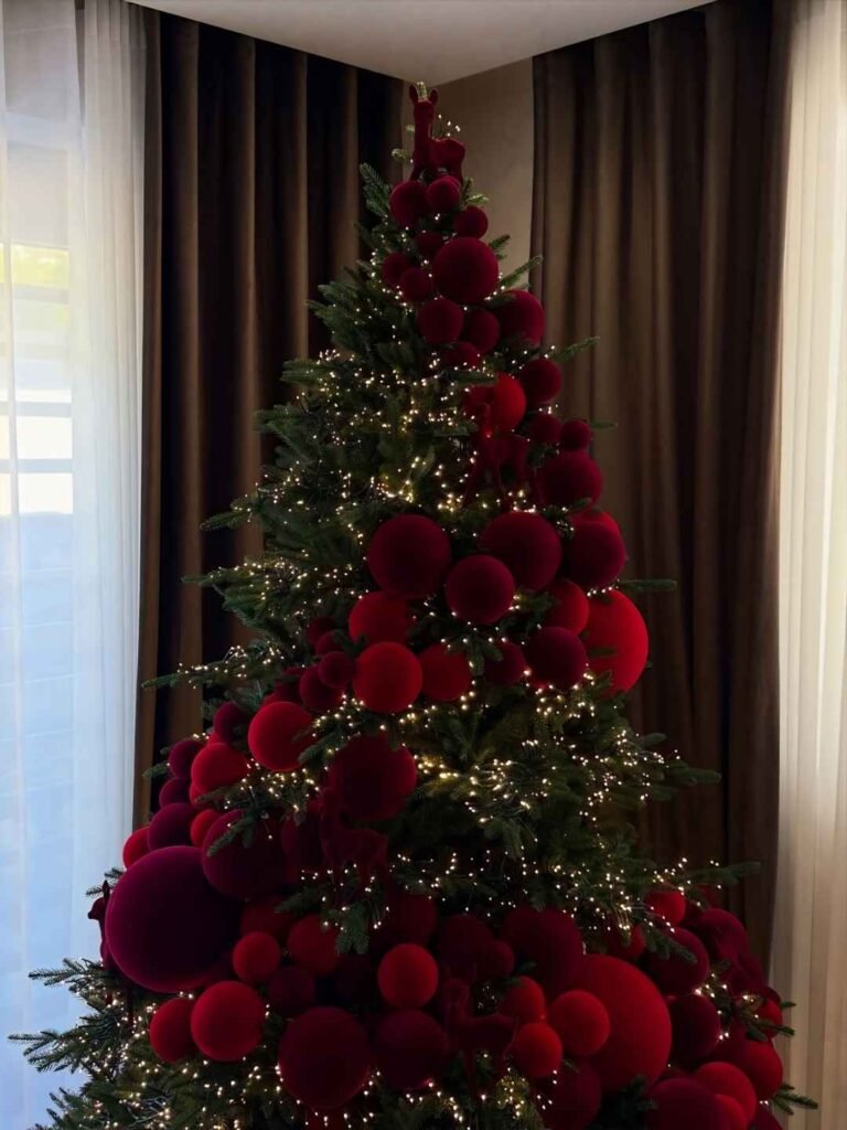 Christmas tree decorated with oversized burgundy and crimson velvet-look baubles, red reindeer figures, and warm white fairy lights.