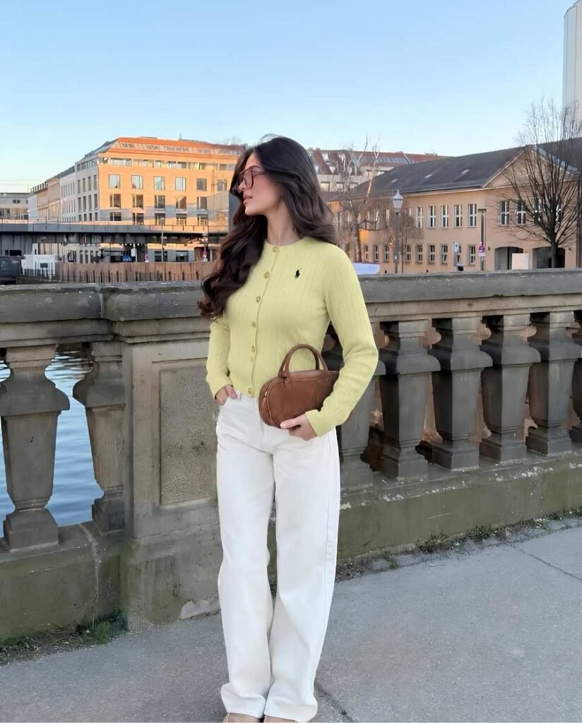 Woman wearing a pastel yellow ribbed button-front cardigan, white wide-leg jeans, brown sunglasses, and a small brown leather handbag beside a stone bridge.