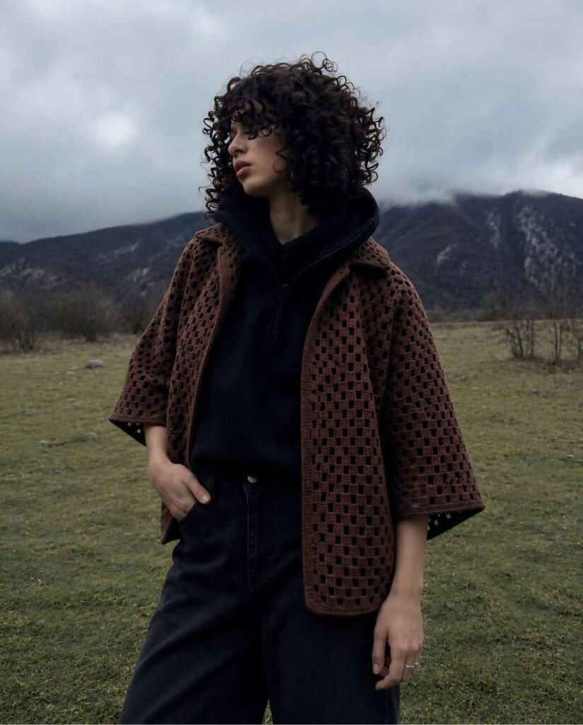 Woman with curly hair wearing a chocolate brown open-knit crochet cardigan over a black zip-up hoodie and loose black jeans in a grassy mountain landscape.