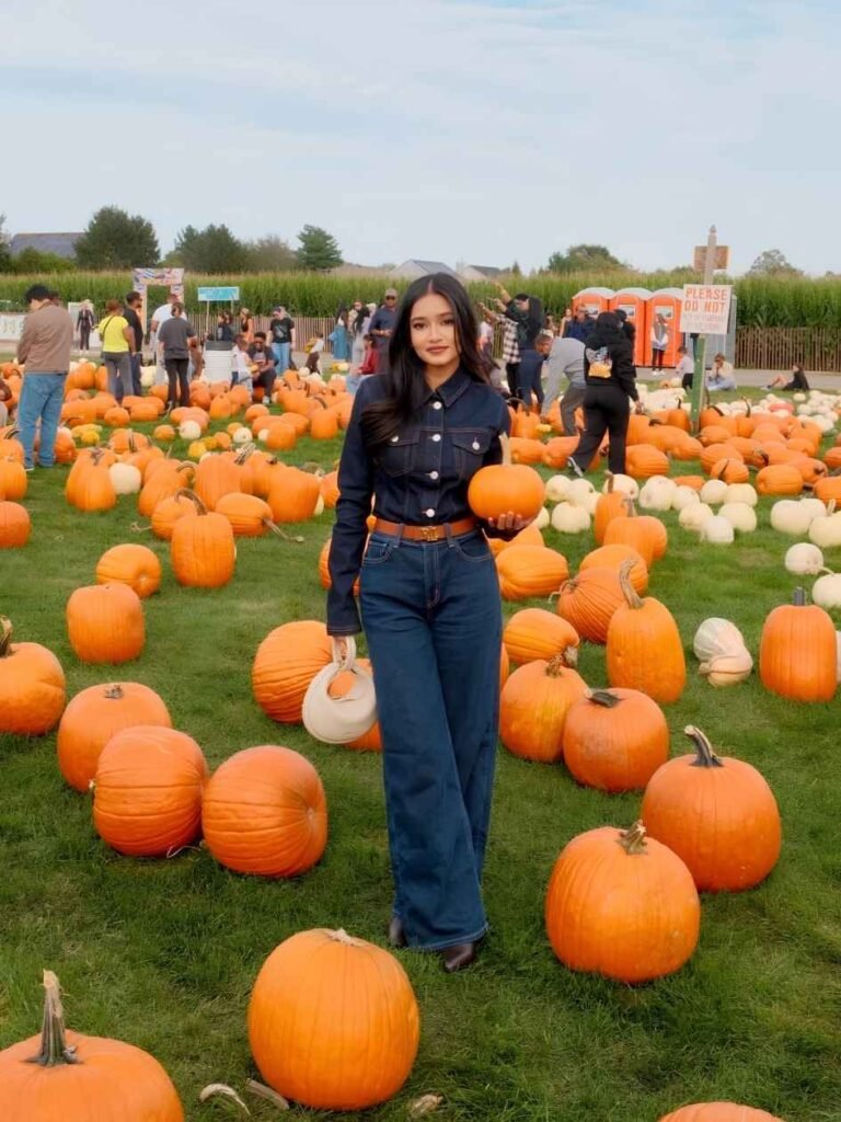 Woman wearing a dark denim button-front shirt, matching wide-leg jeans, brown belt, dark brown boots, and a cream handbag while holding a pumpkin in a pumpkin patch.