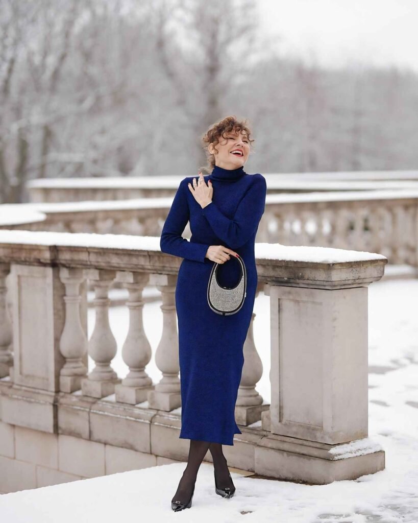 Woman wearing a royal blue long-sleeve turtleneck midi dress, sheer black tights, black pointed-toe heels, and carrying a small silver-embellished handbag outdoors in the snow.