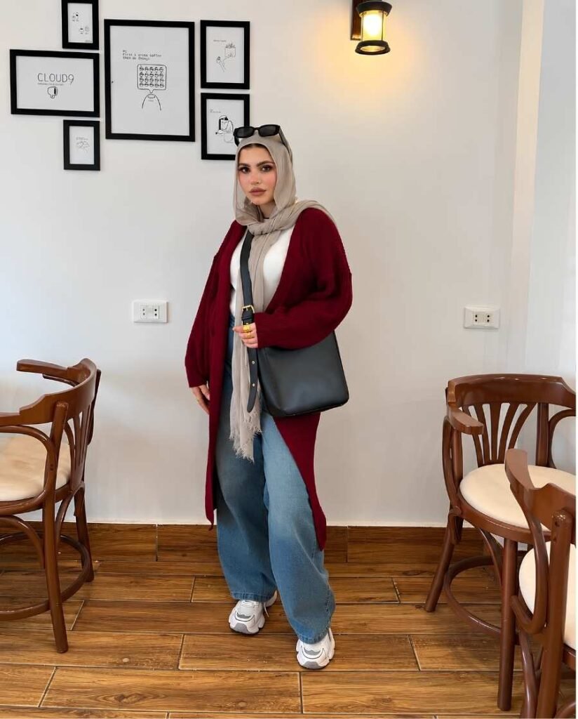 Woman wearing a burgundy longline cardigan, beige hijab, white top, light-blue wide-leg jeans, gray shoulder bag, and chunky sneakers inside a café.
