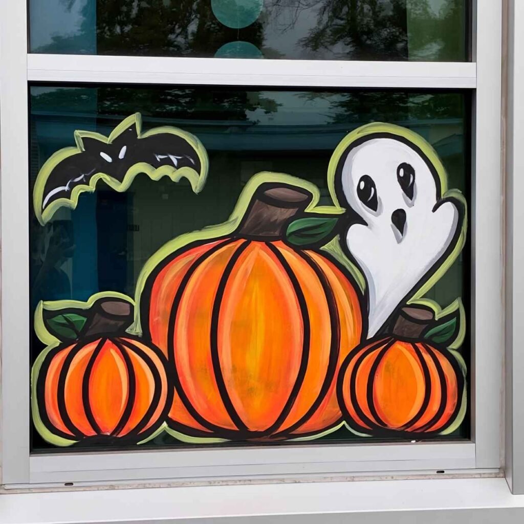 Halloween window painting showing three bright orange pumpkins, a cute white ghost with wide eyes, and a black bat outlined in pale green.