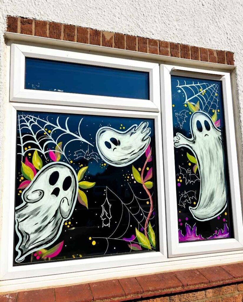 Halloween window painting featuring three floating white ghosts, delicate spiderwebs, small bats, colorful leaves, and yellow dots across two glass window sections.