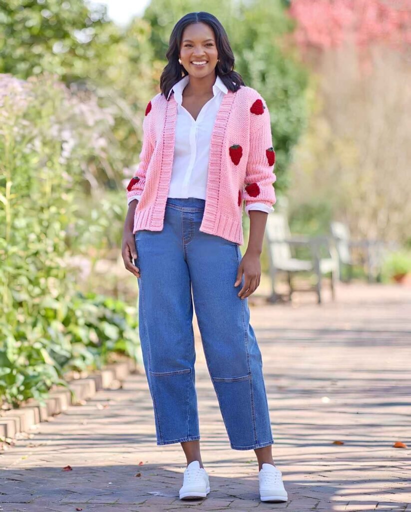  Woman in a pink strawberry-patterned cardigan, white button-down shirt, relaxed blue jeans, and white sneakers standing on a garden walkway.