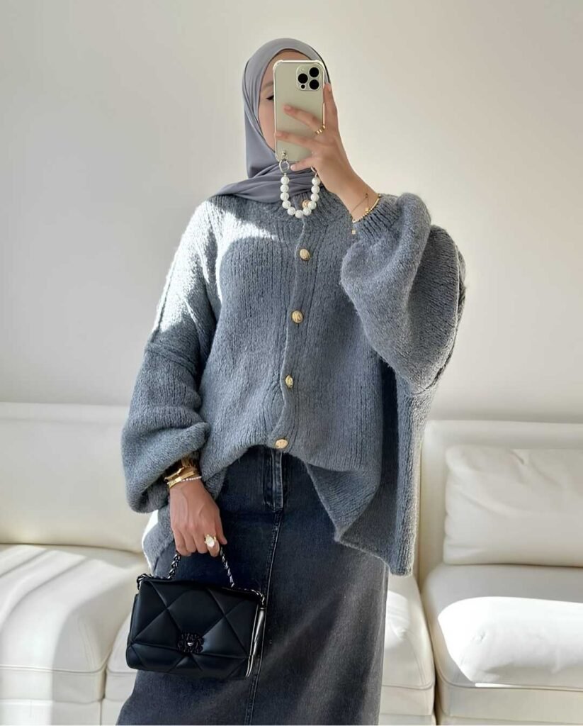 Woman wearing an oversized gray button-front cardigan, charcoal denim maxi skirt, matching gray hijab, and gold jewelry while holding a black quilted handbag.