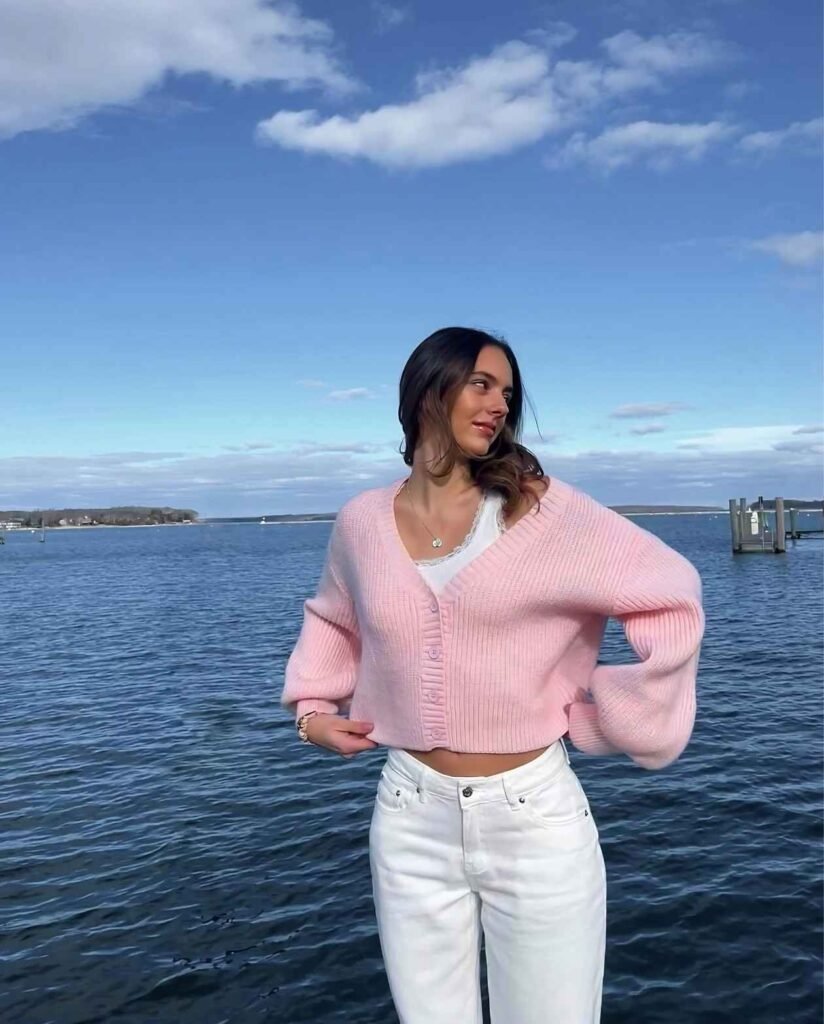Woman wearing a pink cropped ribbed cardigan with balloon sleeves, white lace-trimmed top, and high-waisted white jeans while posing beside a waterfront.