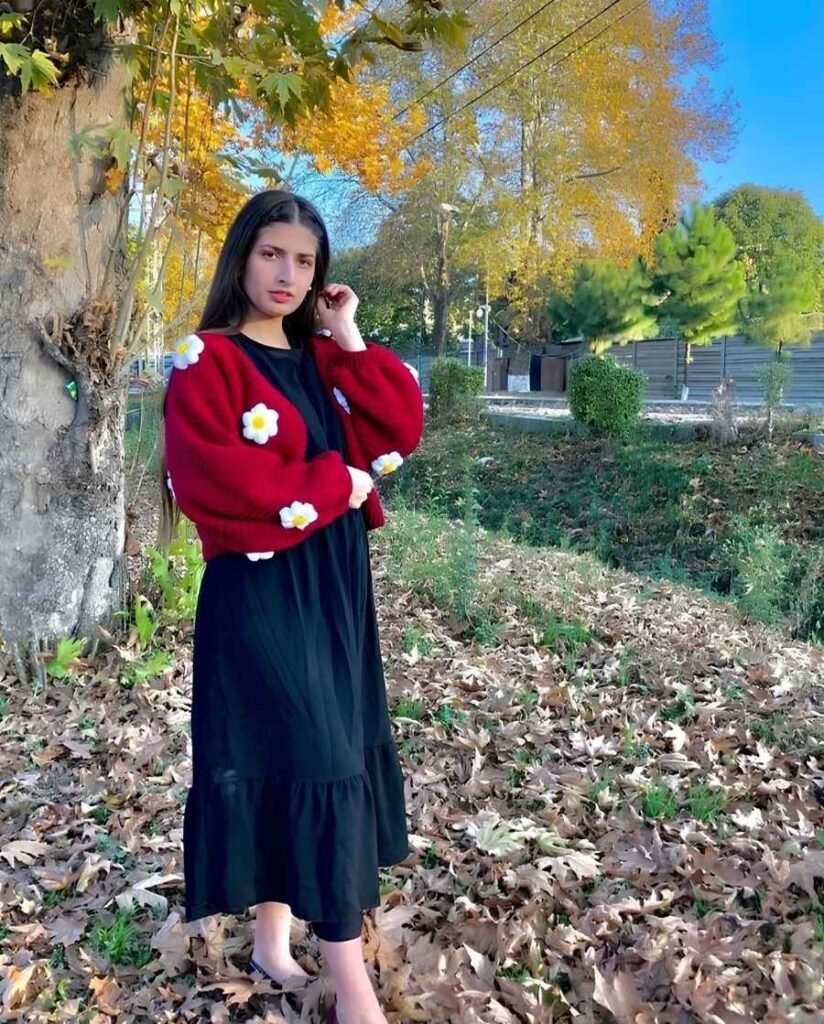 Woman wearing a burgundy chunky-knit cardigan with white daisy appliqués over a flowing black maxi dress and black flats in an autumn garden.