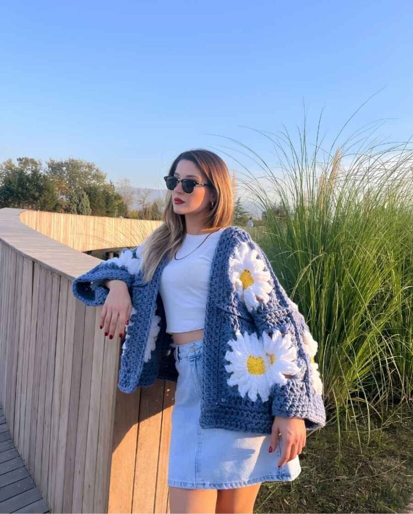 Woman wearing an oversized blue crochet cardigan with white daisy flowers, a white cropped top, light denim mini skirt, and black sunglasses outdoors.