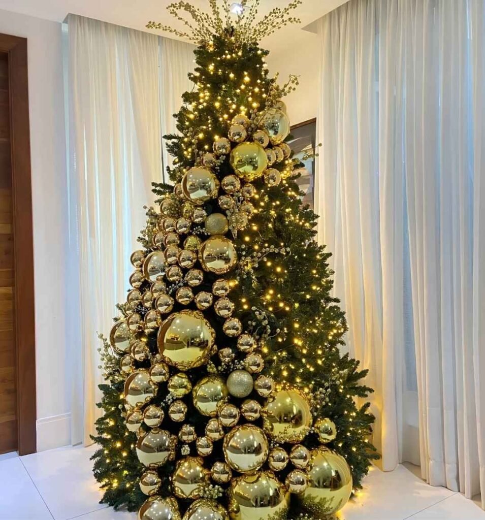Tall Christmas tree with cascading oversized gold baubles, warm white fairy lights, and delicate gold berry sprays in an elegant living room.
