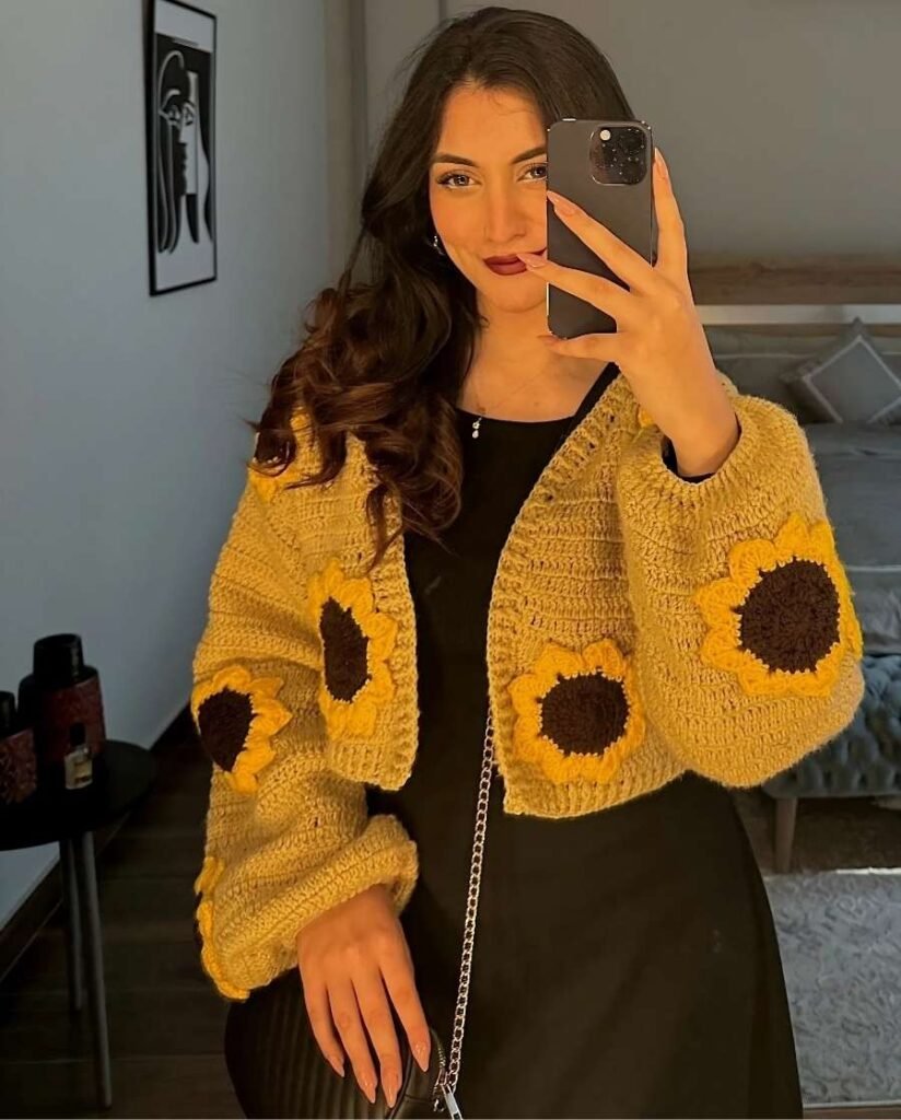 Woman wearing a mustard yellow sunflower crochet cardigan over a fitted black dress, accessorized with a gold-chain crossbody bag and delicate necklace.