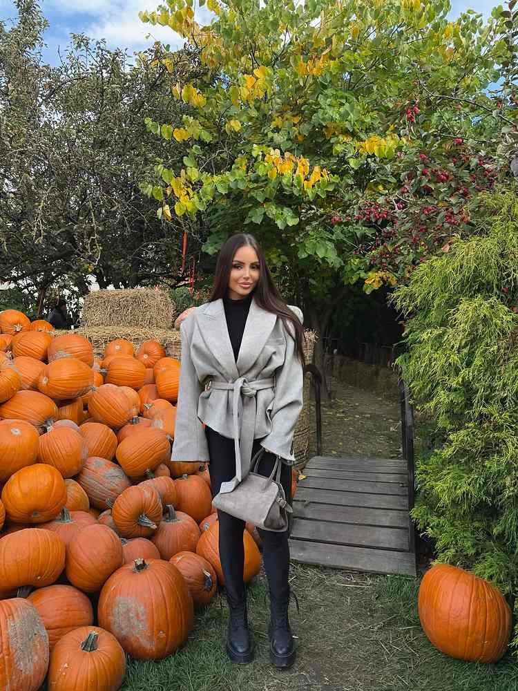 Woman standing beside orange pumpkins wearing a light gray belted oversized blazer, black high-neck top, black leggings, chunky lace-up boots, and a gray handbag.