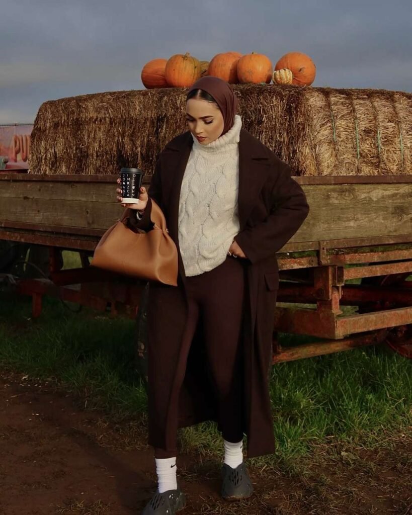 Woman at a pumpkin patch wearing a long chocolate brown coat, cream cable-knit turtleneck sweater, brown fitted pants, matching headscarf, white socks, dark slip-on shoes, and a tan handbag beside hay bales and pumpkins.