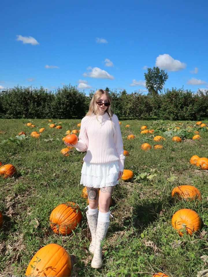 Woman in a pale pink ribbed sweater, white tiered mini skirt, floral knee-high rain boots, and sunglasses holding a small pumpkin in a sunny pumpkin patch.