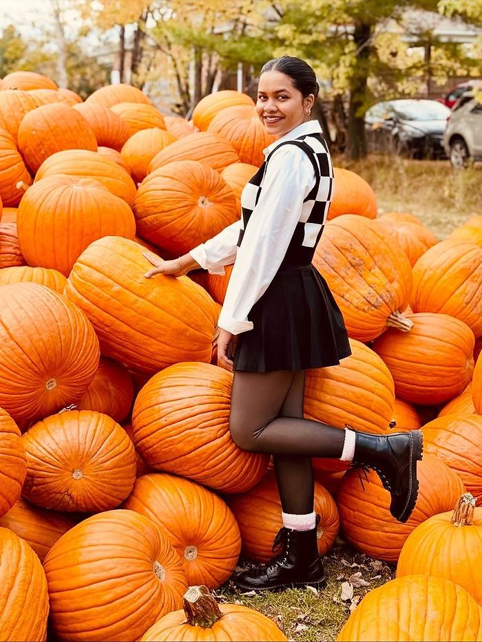 Woman wearing a black-and-white checkered sweater vest over a white button-down shirt, black pleated mini skirt, sheer tights, and black lace-up boots beside a large pile of orange pumpkins.