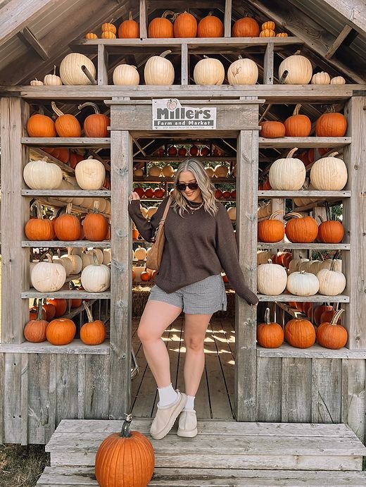 Woman wearing an oversized brown sweater, gray tailored shorts, beige ankle boots, white socks, sunglasses, and a brown shoulder bag outside a rustic pumpkin market.