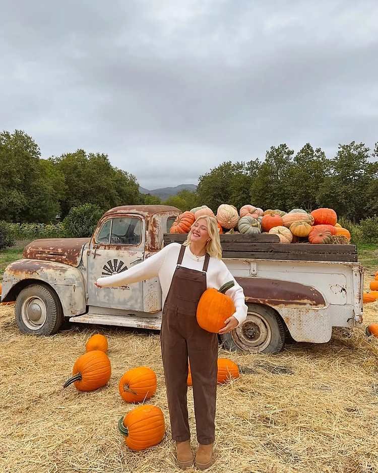 Woman wearing brown bib overalls, a white knit sweater, and tan ankle boots while holding an orange pumpkin in front of a vintage pickup truck filled with pumpkins.