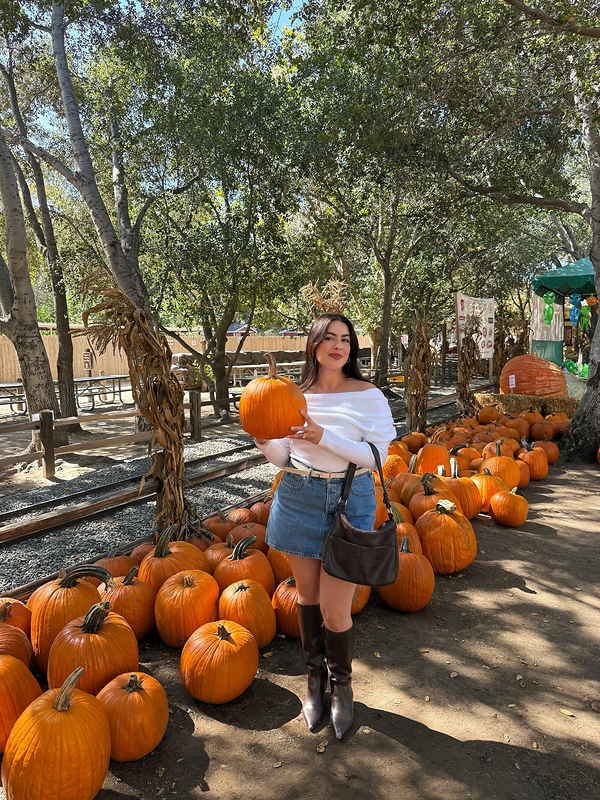 Woman holding an orange pumpkin while wearing a white off-shoulder sweater, blue denim mini skirt, cream belt, dark brown knee-high boots, and a brown shoulder bag at a pumpkin patch.