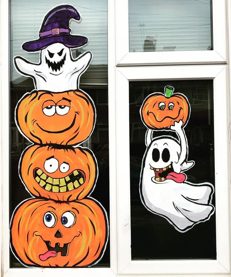 Halloween window painting showing three funny stacked orange pumpkins topped with a purple-hatted ghost, alongside another white ghost holding a small pumpkin.