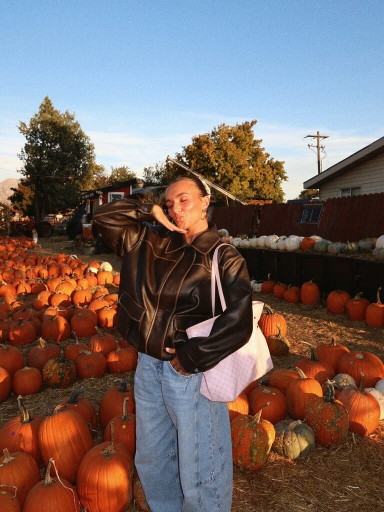 Woman wearing an oversized dark brown leather-look jacket, light blue baggy jeans, gold hoop earrings, and a pale pink shoulder bag at a pumpkin patch.