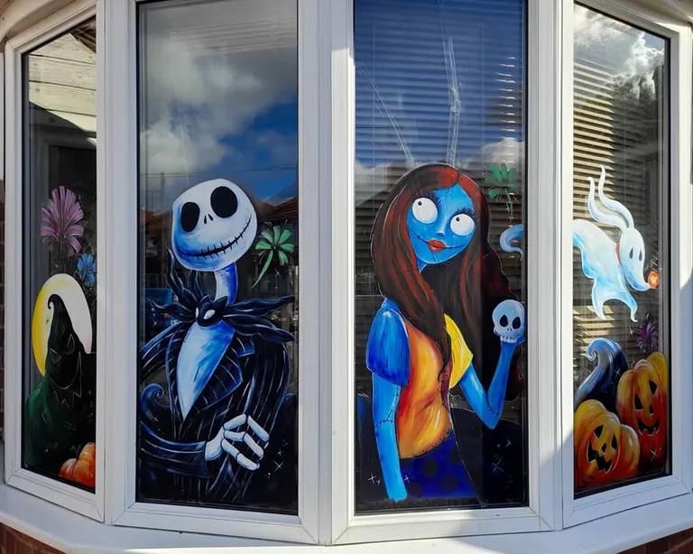 Halloween bay window painting featuring Jack Skellington, Sally holding a skull, Zero the ghost dog, orange pumpkins, and a dark cloaked figure.